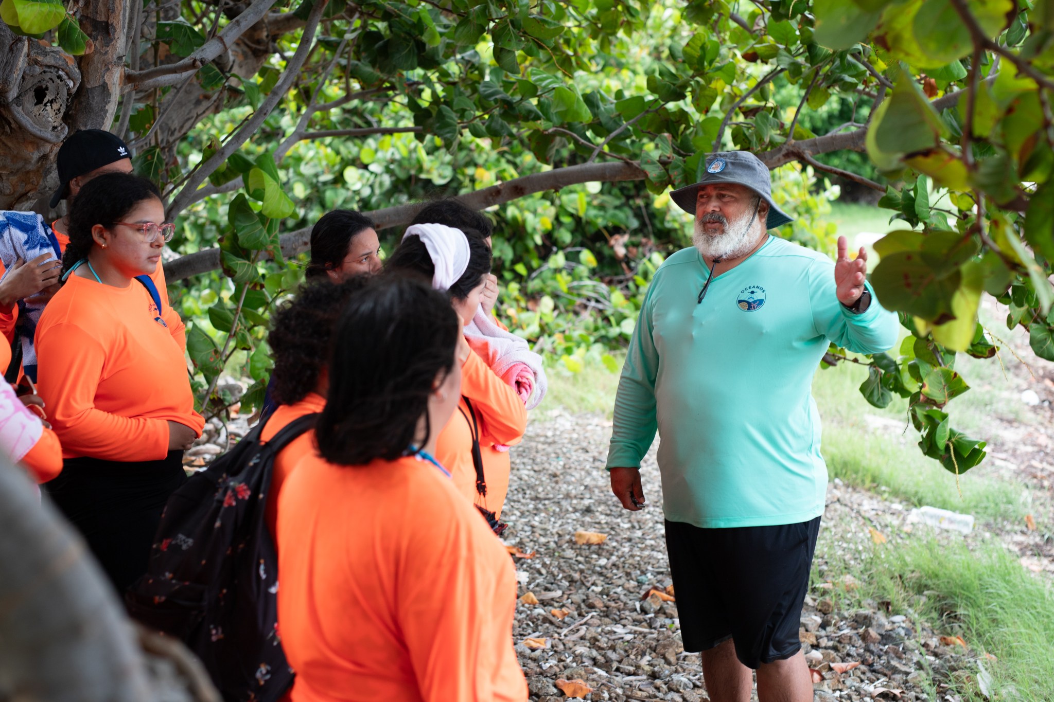 A Puerto Rican man with a bushy white beard, green bucket hat, teal longsleeve shirt, and black shorts gestures to a group of high school students wearing neon orange shirts.