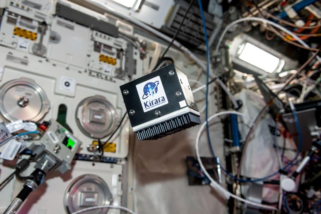 Close-up of a floating black cube-shaped device labeled “Kirara” inside a laboratory filled with cables, equipment, and scientific instruments aboard the International Space Station.