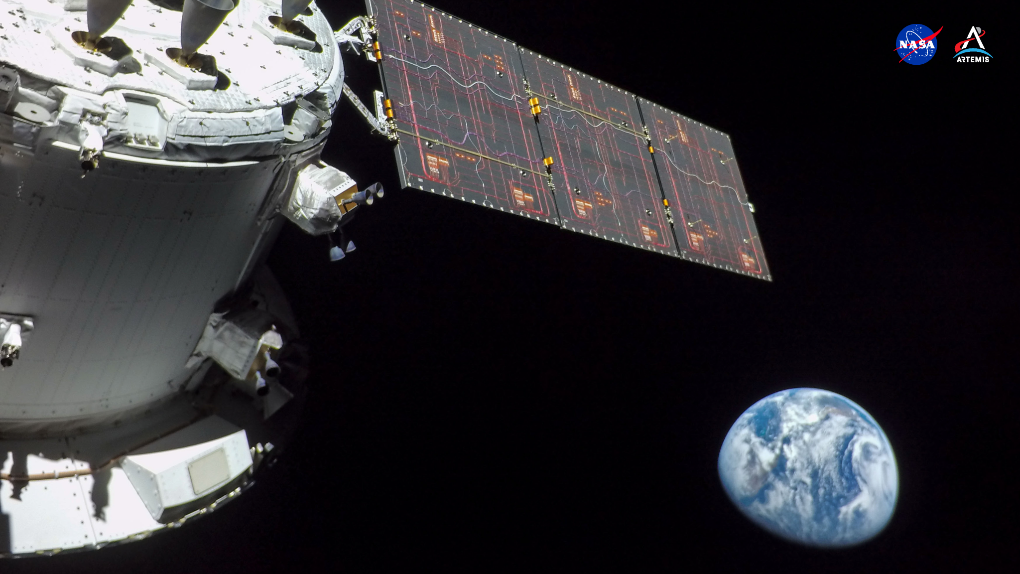 In this first high-resolution image, taken on the first day of the Artemis I mission, the Orion spacecraft’s service module is seen at left, seemingly framing a slightly blurred Earth to its right with one dark, rectangular solar array. Intricate reddish wires cover the solar array like fine threads. The spacecraft, gleaming white, is slightly shadowed against the black expanse of space.