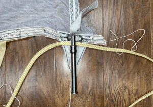 A sample of a Main parachute canopy at the skirt where a reefing line cutter is located. Each parachute has four reefing line cutters, two on each reefing line, to allow the parachute to stage and gradually increase in size during operation. The pyrotechnic cutter seen in the middle is about the size of a permanent marker and contains pyrotechnics that operate as a timing fuse and output charge to propel a blade through the Kevlar reefing line seen lying below it in this photo. Credit: Courtney Beasley/Jared Daum