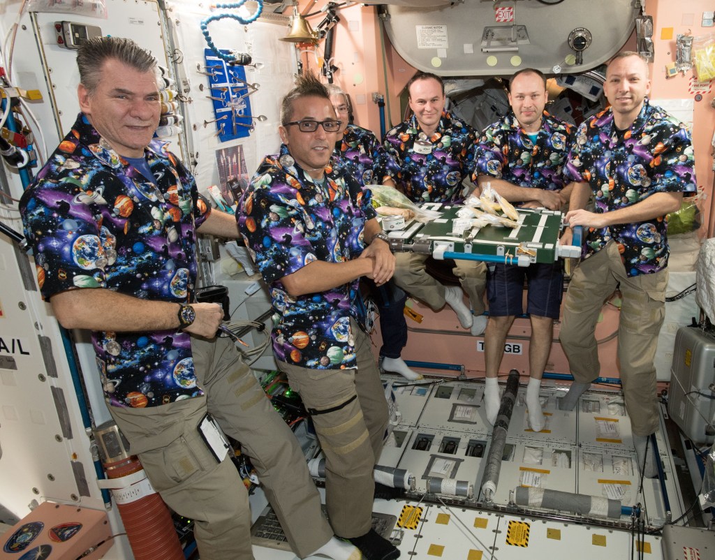 Six astronauts in matching colorful button down shirts with planets, stars, and spacecrafts on them in purple, orange, blues, and green are all floating around a small table. The table has food on it. They are onboard NASA's space station. The back wall is pink and the left wall and floors are light grey. Four of the astronauts are wearing beige pants, one is wearing blue shorts, and one is wearing black pants. They are all smiling into the camera.