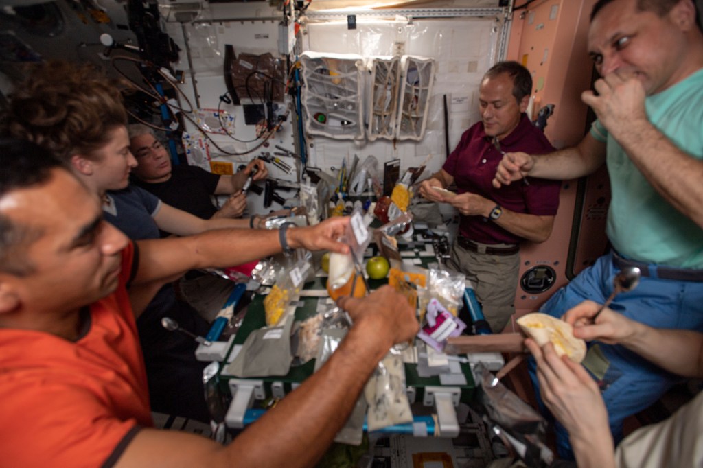 Five astronauts, four men and one woman, are surrounding a table covered in bags of food. A pair of hands belonging to someone whose face is offscreen is holding a tortilla and a spoon. They are all focusing on what is in front of them.