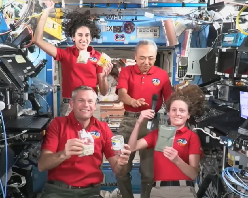 Four astronauts in red shirts are smiling into the camera. They are holding various food items. Some of the food items are floating. One astronaut that is a man is standing in the back right of the image. He is reaching to grab the black microphone that is in the hand of the woman in front of him.