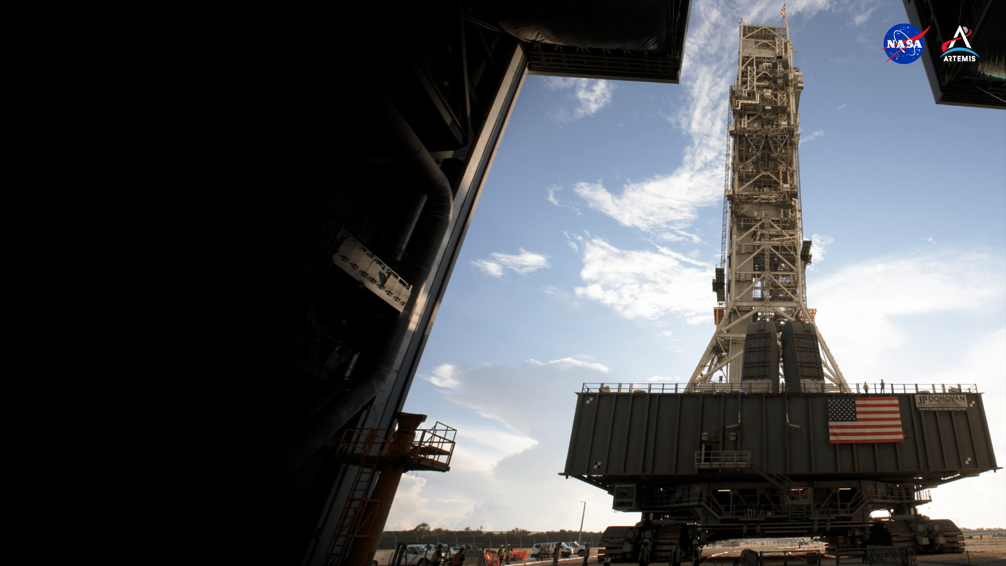 Framed by a blue sky and white clouds, NASA’s mobile launcher — a vertical, rectangular structure constructed of gray metal and open scaffolding — seemingly rises into the sky. At the top of the structure is an American flag waving in the wind. The mobile launcher sits atop Crawler Transporter-2 — a flat, square platform that carries heavy structures to the launch pad on tooth-metal wheels, or sprockets, which are commonly seen on tanks. Crawler Transporter-2 also has an American flag affixed to the front, which lies flat against the structure.