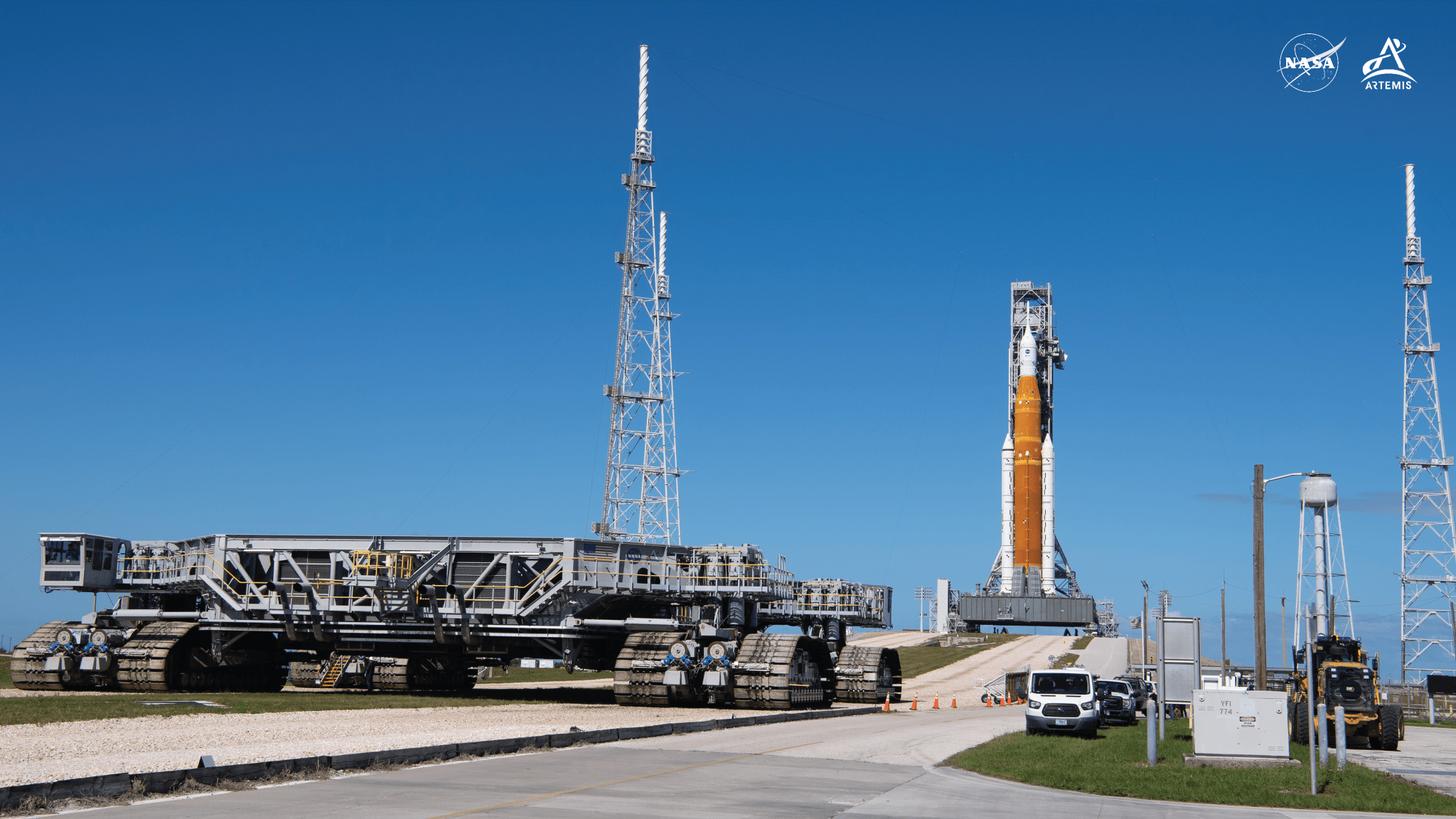 Backdropped by a nearly cloudless blue sky, Crawler Transporter-2 — a flat, square platform that carries heavy structures to the launch pad on tooth-metal wheels, or sprockets, which are commonly seen on tanks — dominates the foreground of the image as it moves away from the launch pad. In the background near the right-hand side of the image, and in between two metal tower structures and one metal water tower, NASA’s SLS rocket with Orion spacecraft aboard is seen connected to and in front of the mobile launcher at Launch Pad 39B on Nov. 4, 2022. The orange core stage of the SLS rocket stands out from the white solid rocket boosters at each side and white Interim Cryogenic Propulsion Stage and white Orion capsule topping it.