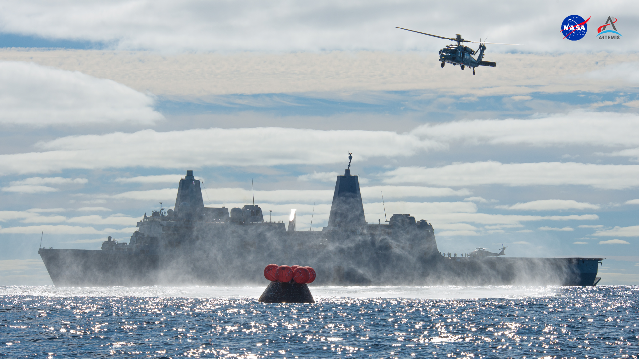 In this image, at the foreground and in front of the massive USS Portland aircraft carrier ship, is the dark gray Orion capsule bobbing in the ocean after returning from its flight test around the Moon. Directly above the capsule are five visible inflated orange balloons. A large Navy helicopter is seen in a cloud-filled sky as it approaches the spacecraft from overhead.