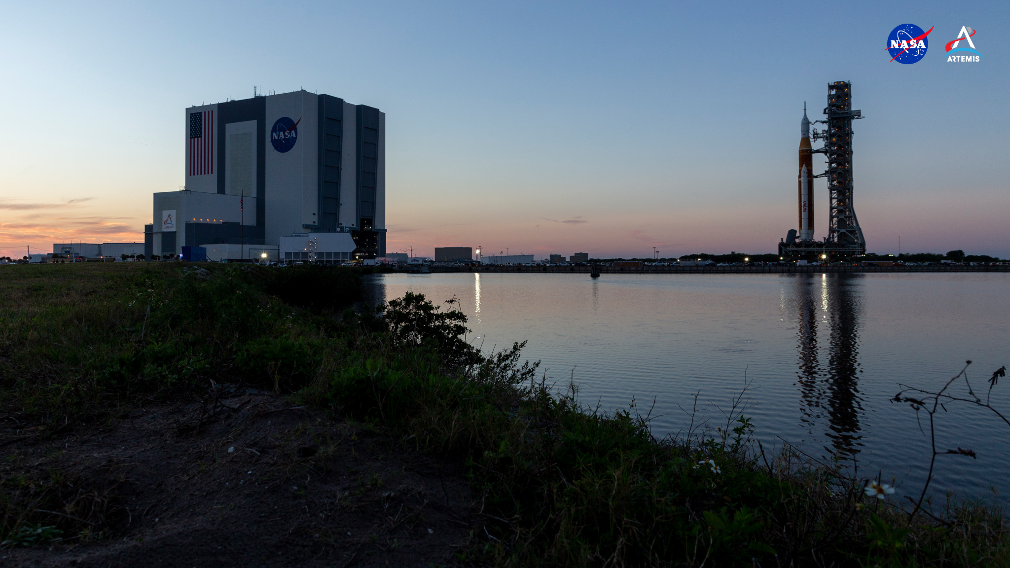 Backdropped against the pastel hues of a dawn sky, at left in the image is the Vehicle Assembly Building, a square, windowless building with gray accents and an enormous, painted American flag next to another painted NASA insignia, nicknamed the “meatball.” At right, the towering, interconnected structures of NASA’s crawler transporter, mobile launcher, and SLS rocket with Orion spacecraft atop roll out toward Launch Complex 39B for the first time on March 17, 2022, at NASA’s Kennedy Space Center in Florida. The scene is framed by shadowed vegetation and a large body of water in the foreground.