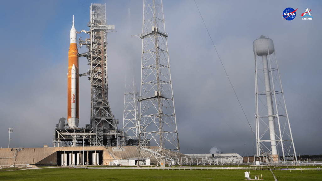 In this close-up image, NASA’s SLS rocket with Orion spacecraft aboard is seen atop the mobile launcher at Launch Pad 39B on Nov. 12, 2022, at NASA’s Kennedy Space Center in Florida. Slightly off center and at left, the rocket rises above the massive concrete launch pad, its solid rocket booster, upper stage, and Orion spacecraft standing out in white against the sturdy orange core stage. Metallic arms from the rectangular scaffolding of the mobile launcher extend to hold the rocket in place. Also visible within the mist of fog at the launch pad are other metal radio tower structures and a water tower.