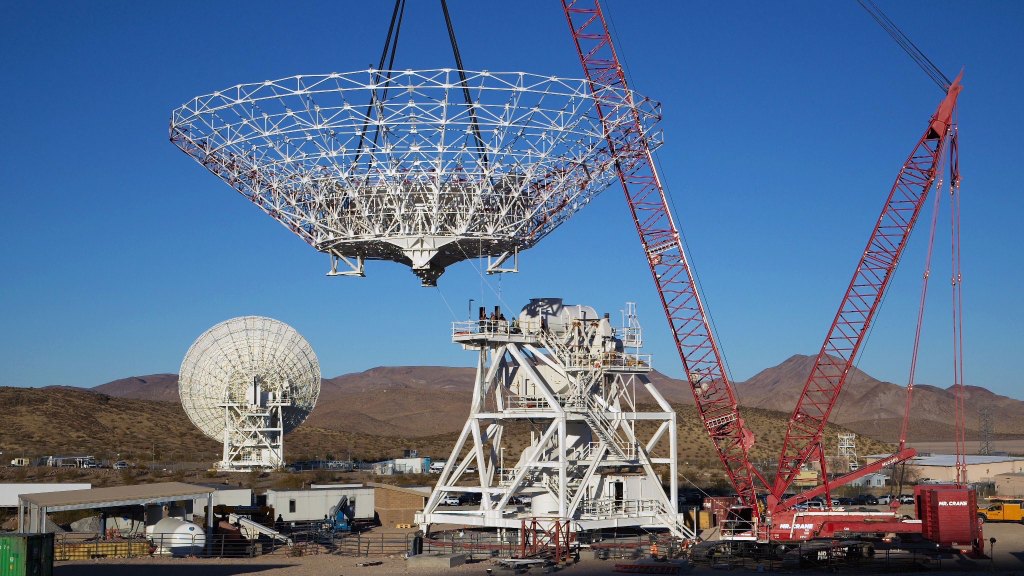 A large cranes lifts a massive white dish antenna into place against a clear blue sky. The antenna's intricate framework is visible as it is carefully positioned. In the background, a smaller antenna and desert hills complete the scene.