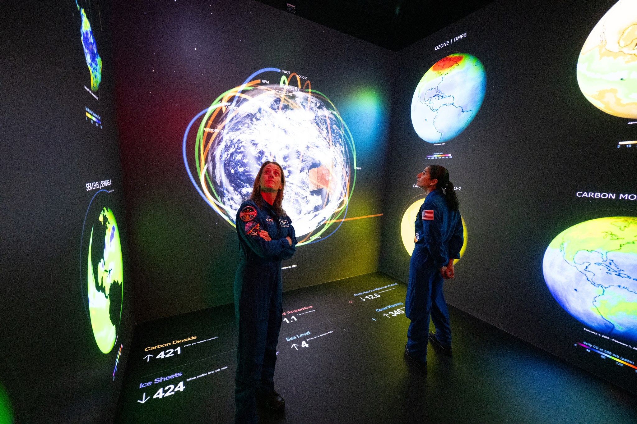 Two women in blue jumpsuits stand in a room with black walls and a black floor. The walls and floor have infographics, and data points on them related to Earth. The woman at left, NASA astronaut Loral O'Hara, looks up and to the side. The other woman, NASA astronaut Jasmin Moghbeli, faces the other way.