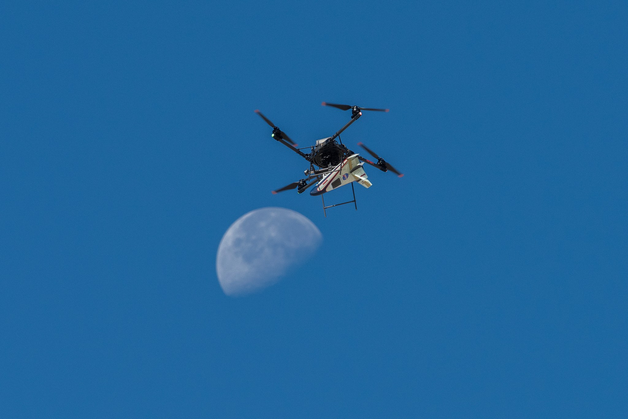 A white, blue, and red probe attached to a rotor with four blades flies in the blue sky, just above the Moon.