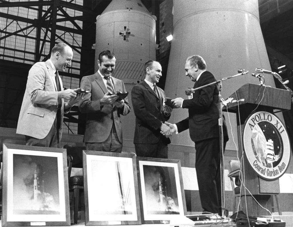 Black and white image of four men in a large hall with spacecraft modules behind them