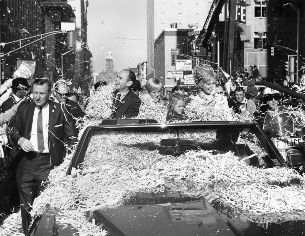 Black and white street level scene of a parade with streamers covering the car and its occupants.