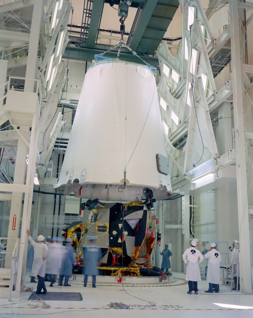 Several men standing around a large room as a large spacecraft component shaped like a truncated cone hangs from a crane.