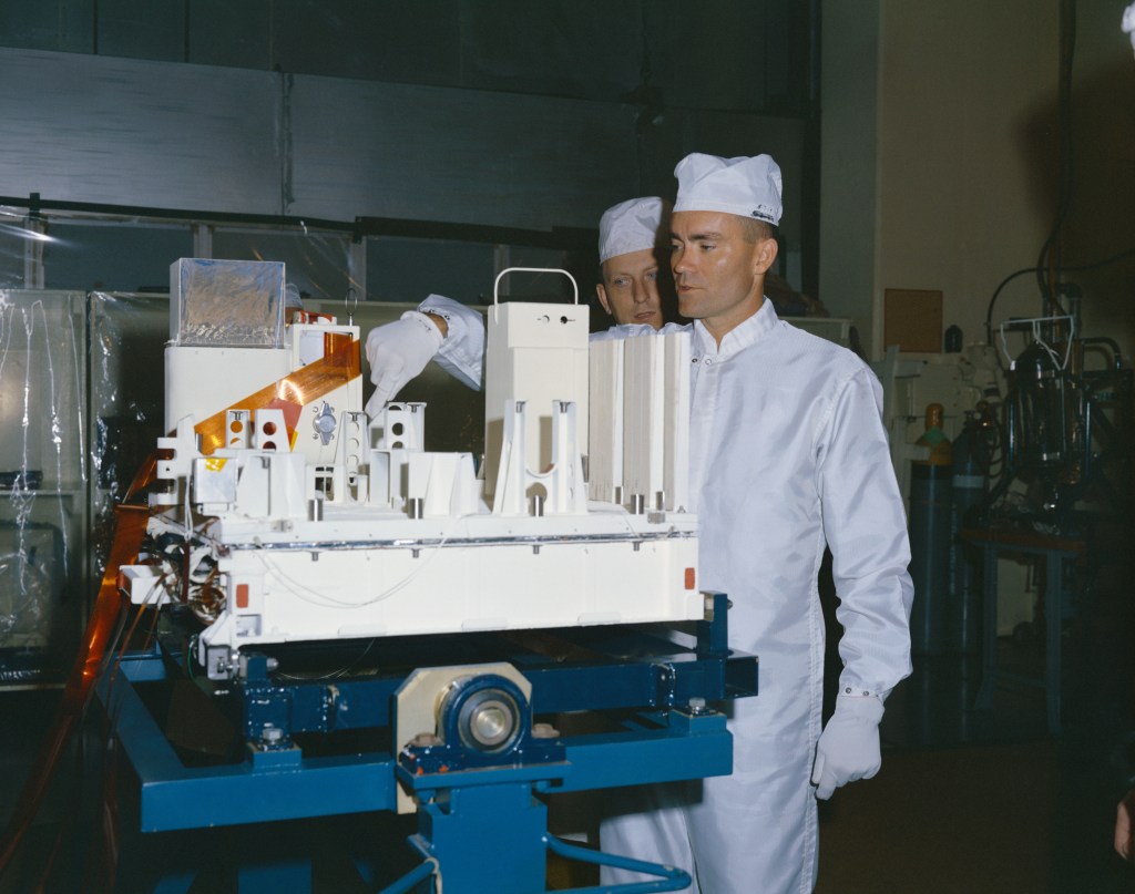 A man dressed in white overalls and a white cap stands next to a scientific instrument