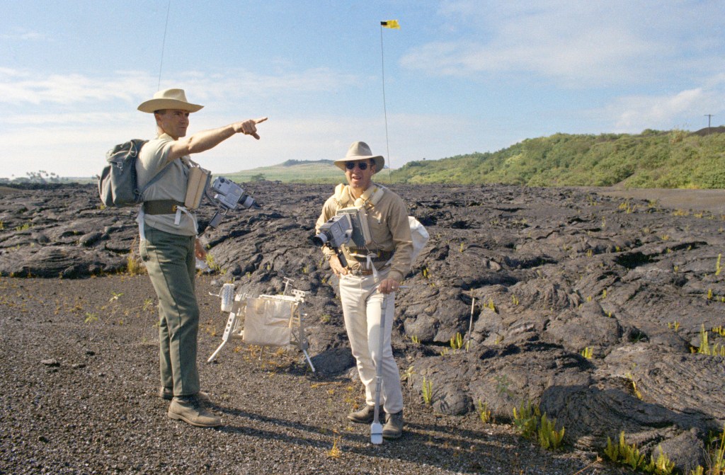 Two men standing over a rugged outdoor terrain, looks like a lava field