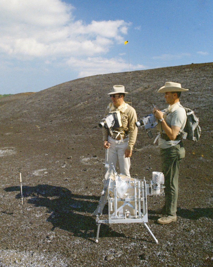 Two men standing on rough ground next to an instrument, with a hill in the background.