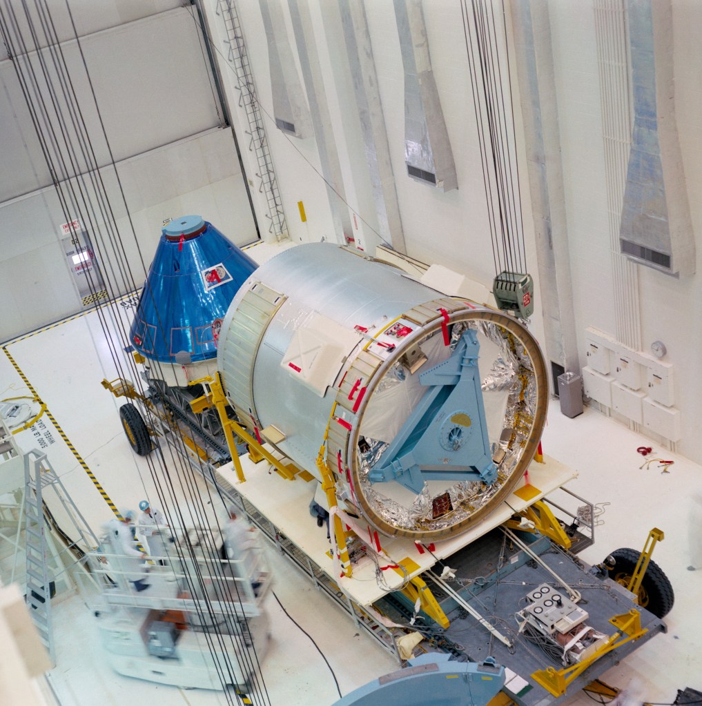 Overhead view of a cylindrical and a conical spacecraft in a large hanger