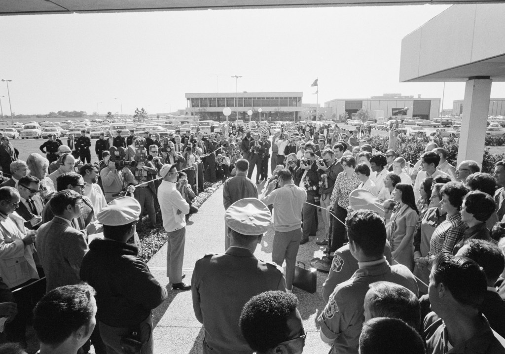 Black and white image of a group of men and women gathered around three men at a microphone.