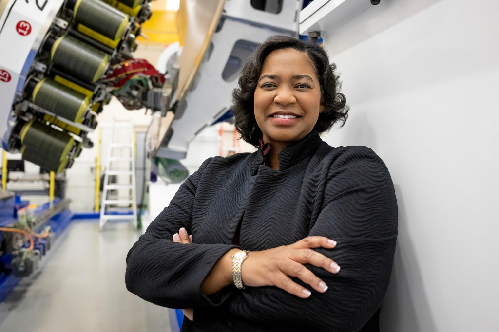 Tawnya Laughinghouse is standing in a high-tech industrial facility, smiling and crossing her arms. She is wearing a textured black jacket and a gold bracelet. Behind her are large pieces of machinery and industrial equipment, with bright lighting highlighting the space.