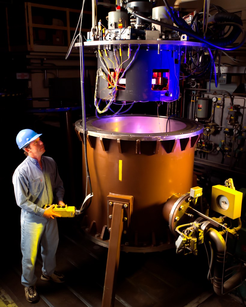 A man wearing a blue hard hat stands next to a large cylinder-shaped rig while operating it.