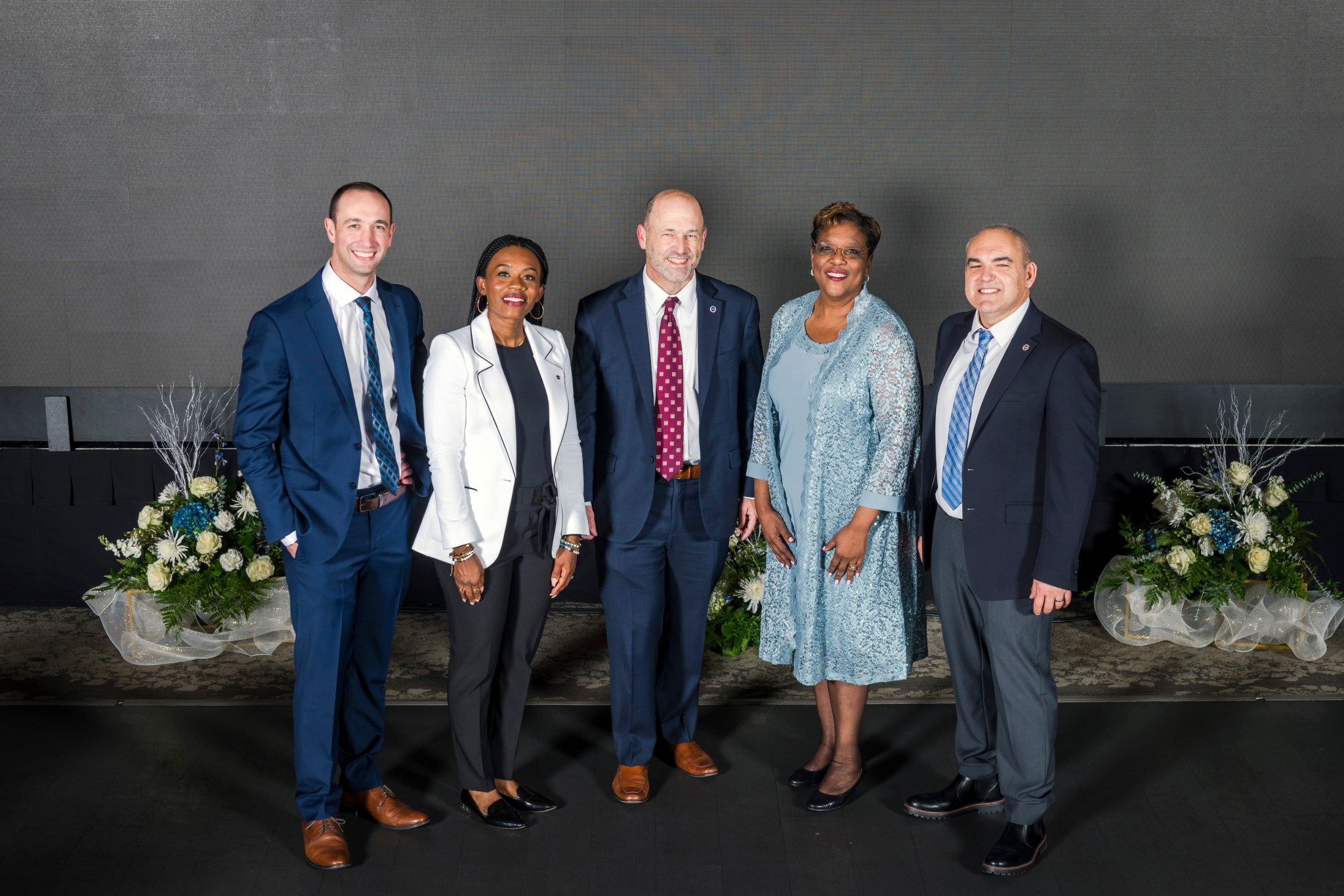 NASA’s Glenn Research Center leaders stand with Evening With the Stars presenters. The group of five people smile at the camera in front of several floral arrangements.