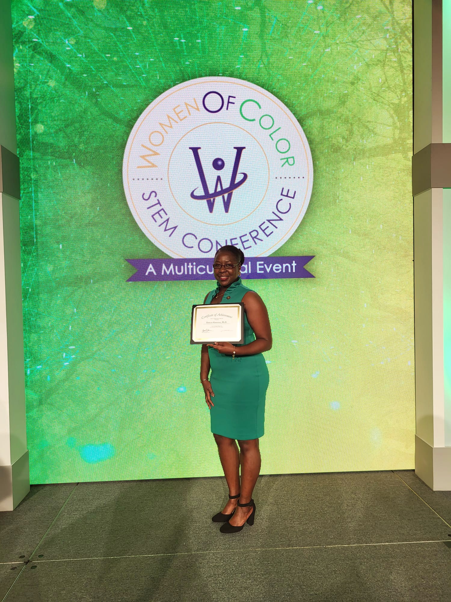 Dorcas Kaweesa A recipient of the Women of Color STEM Conference award is seen standing on stage, holding a certificate of recognition. She is dressed in a green dress and black heels, with the conference logo displayed prominently on the backdrop.