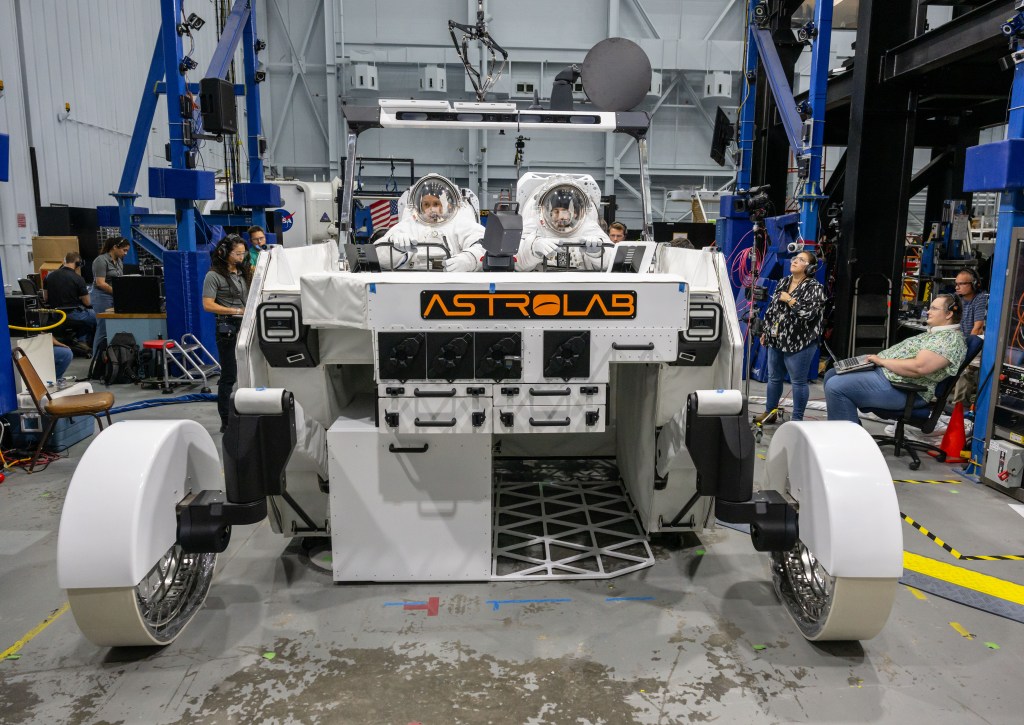 NASA astronaut Frank Rubio and NASA spacesuit engineer Zach Tejral sit inside Astrolab's FLEX lunar terrain vehicle while wearing white spacesuits. The rover is white with an orange Astrolab logo on the front.