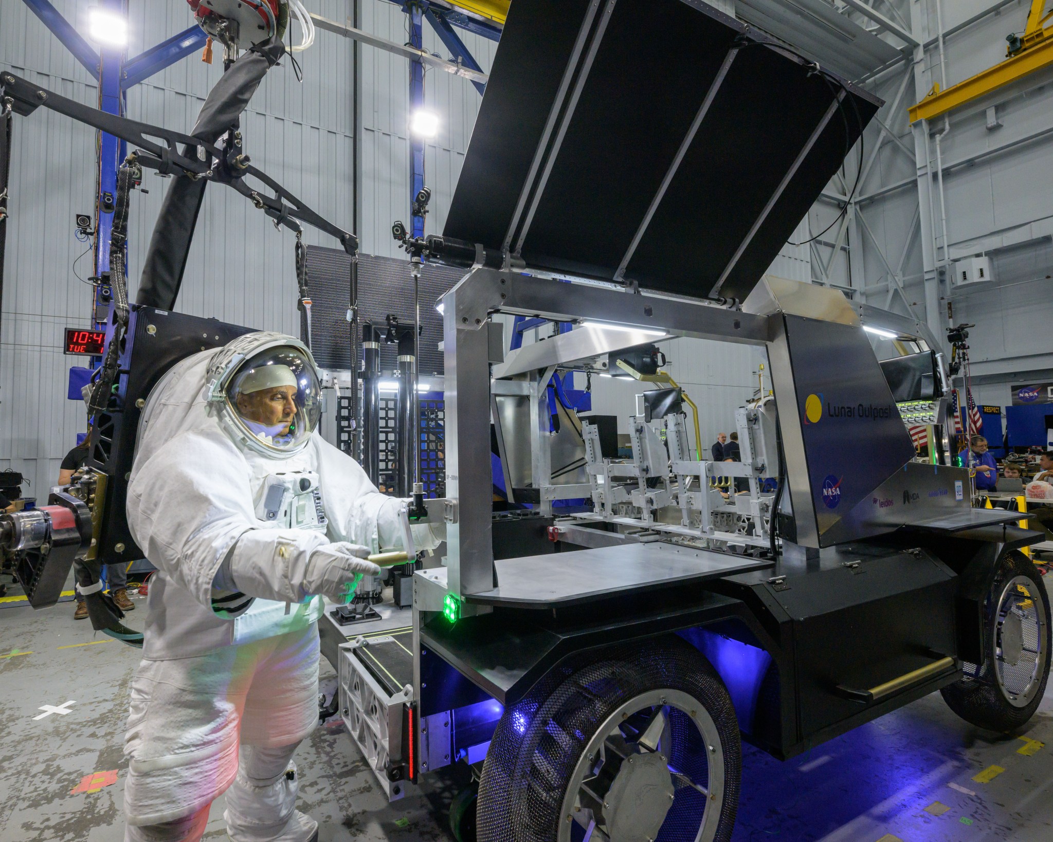 NASA astronaut Joe Acaba is shown in a white spacesuit, rotating a handle to raise a black solar array panel on Lunar Outpost’s Eagle lunar terrain vehicle.