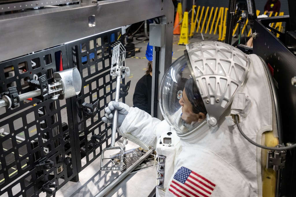 NASA astronaut Jessica Meir is shown in a white spacesuit with the American flag on her left shoulder as she grabs a lunar geology tool from a tool rack on Lunar Outpost's Eagle lunar terrain vehicle.