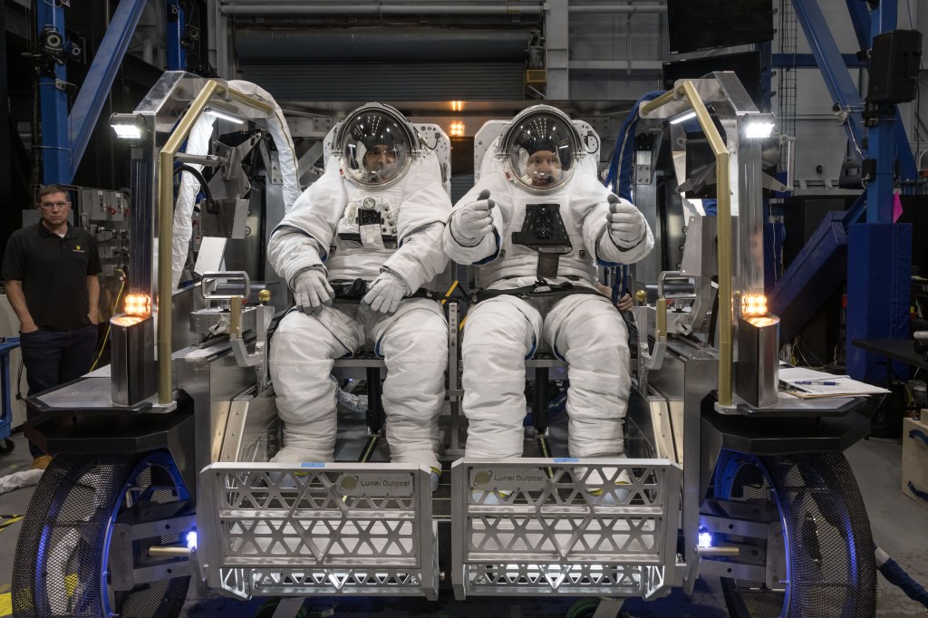 NASA astronauts Raja Chari and Randy Bresnik sit side by side inside Lunar Outpost's Eagle lunar terrain vehicle while wearing white spacesuits.