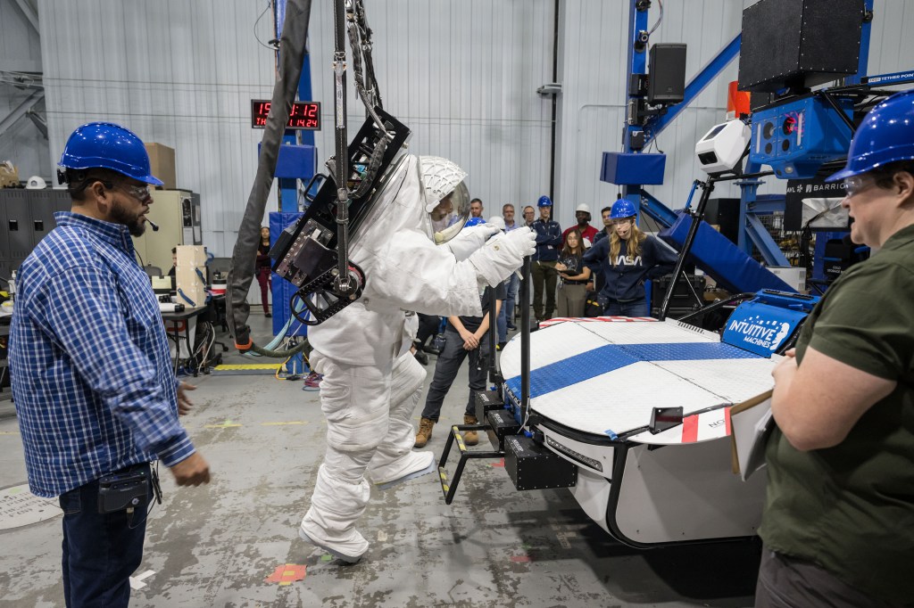 NASA astronaut Joe Acaba is wearing a white spacesuit that is being held up by a crane while he prepares to climb on top of Intuitive Machines' Moon RACER lunar terrain vehicle to get to a black science payload box. The rover is white with a blue stripe down the middle.