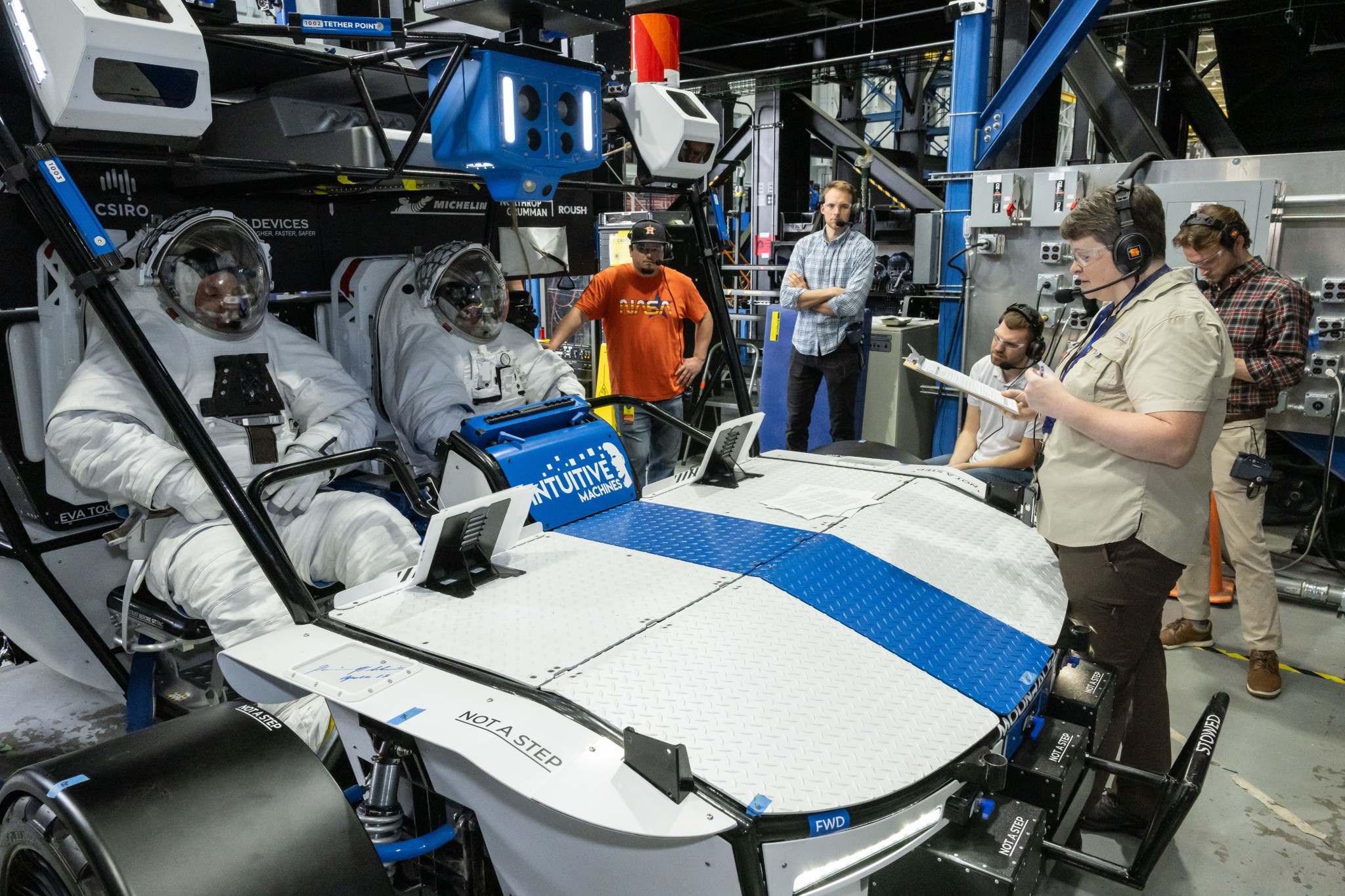 NASA engineer Dave Coan and NASA astronaut Jessica Watkins are wearing white spacesuits while sitting inside of Intuitive Machines’ Moon RACER lunar terrain vehicle. The rover is white with a blue stripe down the middle. There is a test team surrounding them with the test lead is holding a clipboard.