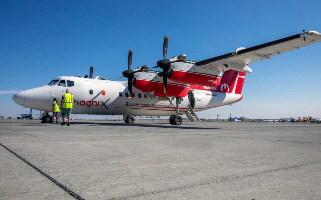 A turboprop plane with a red and white livery sits on a tarmac with a bright blue sky in the background. A man and a woman wearing safety vests stand at the front of the plane. 