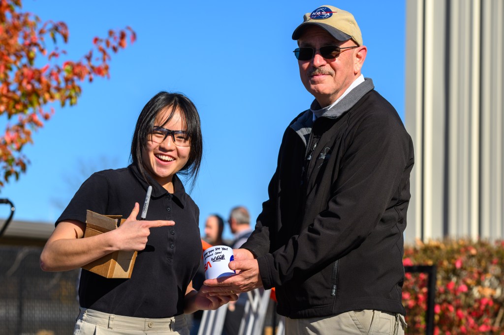 Portage School of Leaders High School junior Rebecca Anderson, wearing a black short-sleeved shirt and glasses, stands smiling beside NASA Glenn’s Daniel Sutliff, an acoustic engineer, on the campus of the University of Notre Dame. Sutliff is wearing a NASA ballcap and black jacket. He and Anderson are holding an autographed NASA mug, which Sutliff signed with a black ink permanent magic marker. Anderson is holding the black marker and a small cardboard box.