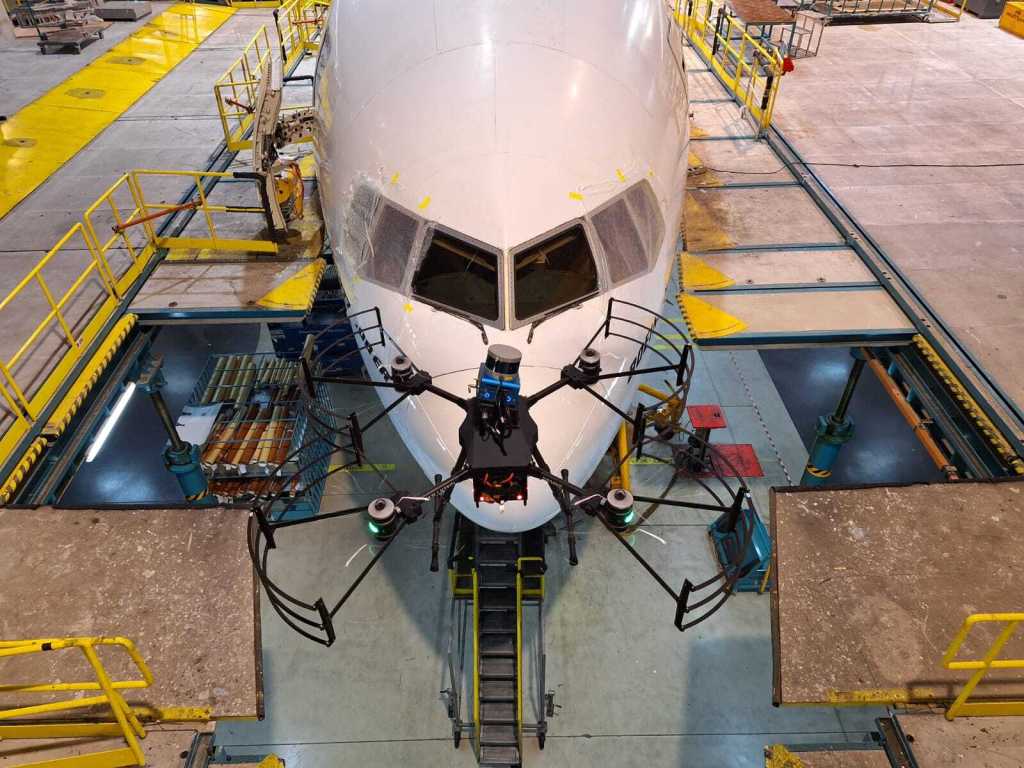 A small, black drone with four rotors is shown in the foreground flying in front of a commercial airliner in the background. The airliner is painted white and the front facing windows can be seen behind the drone. Concrete platforms surround the commercial airliner and yellow ramps connect the platforms to the plane.