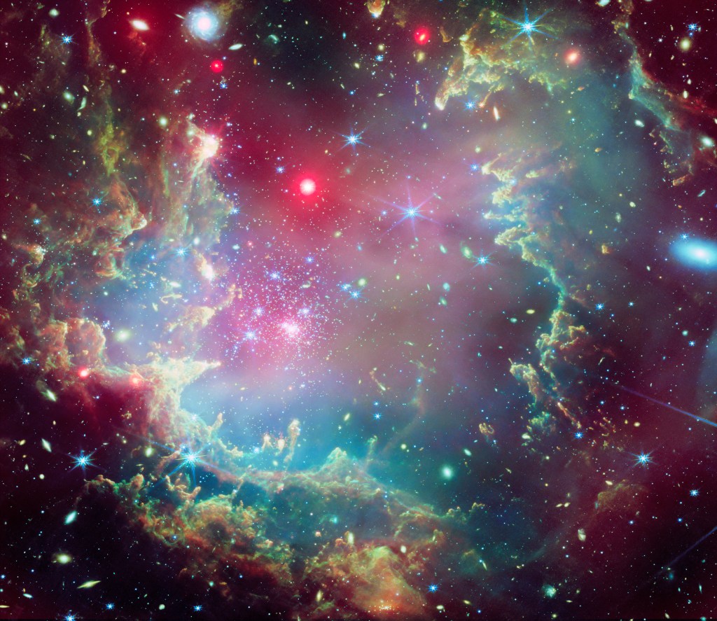 Near the outskirts of the Small Magellanic Cloud, a satellite galaxy roughly 200 000 light-years from Earth, lies the young star cluster NGC 602, which is featured in this new image from the NASA/ESA/CSA James Webb Space Telescope. This image includes data from Webb’s NIRCam (Near-InfraRed Camera) and MIRI (Mid-InfraRed Instrument). The local environment of this cluster is a close analogue of what existed in the early Universe, with very low abundances of elements heavier than hydrogen and helium. The existence of dark clouds of dense dust and the fact that the cluster is rich in ionised gas also suggest the presence of ongoing star formation processes. This cluster provides a valuable opportunity to examine star formation scenarios under dramatically different conditions from those in the solar neighbourhood. [Image description: A star cluster is shown inside a large nebula of many-coloured gas and dust. The material forms dark ridges and peaks of gas and dust surrounding the cluster, lit on the inner side, while layers of diffuse, translucent clouds blanket over them. Around and within the gas, a huge number of distant galaxies can be seen, some quite large, as well as a few stars nearer to us which are very large and bright.]