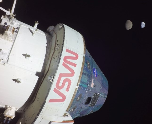 The white Orion capsule with the red NASA logo floats in space against a black background. The Moon and Earth are in the background to the right of the capsule. The Earth is smaller than the Moon. The Moon sits to the left of the Earth.