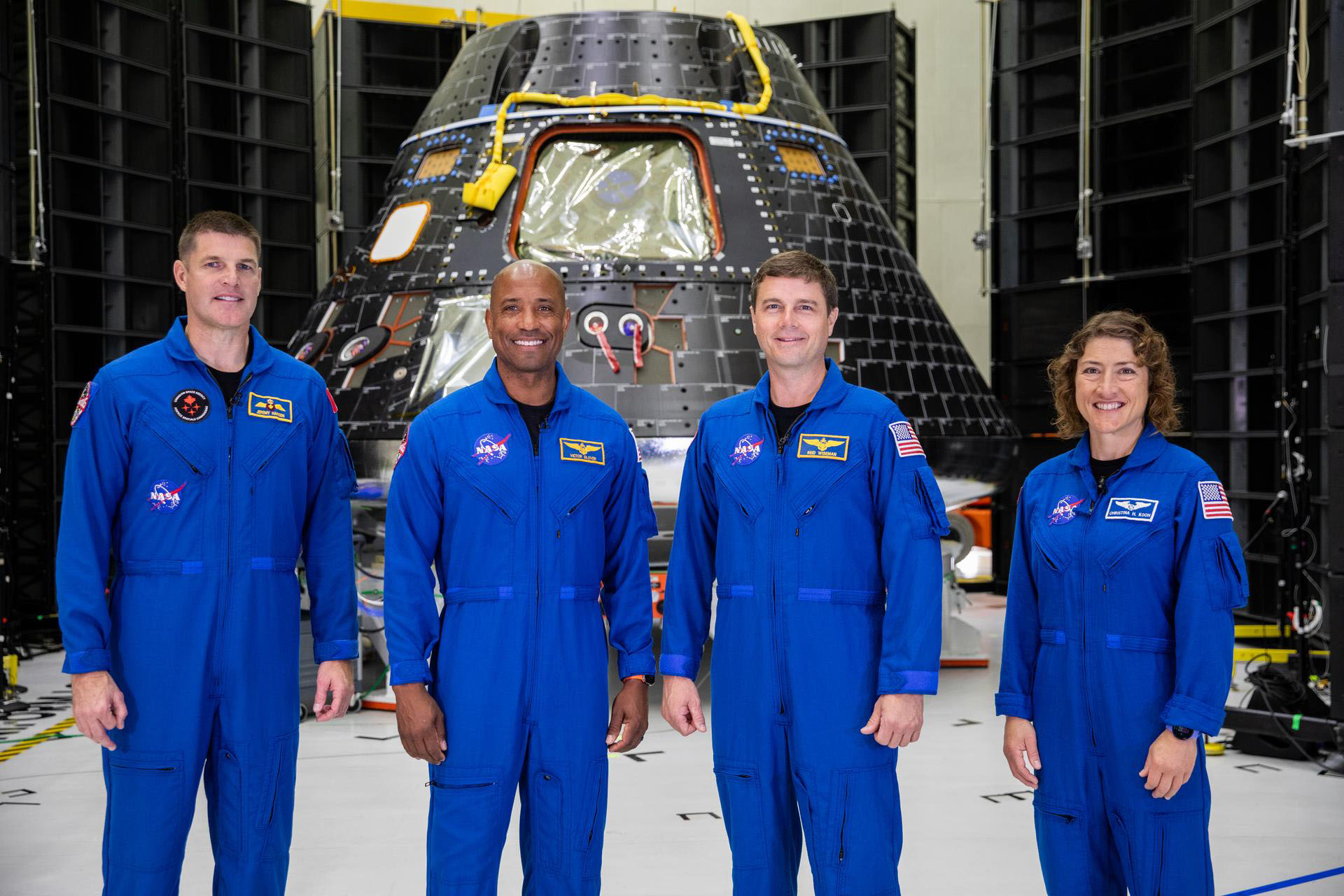 Four crew members in blue jumpsuits are standing next to each other in front of the Orion capsule.