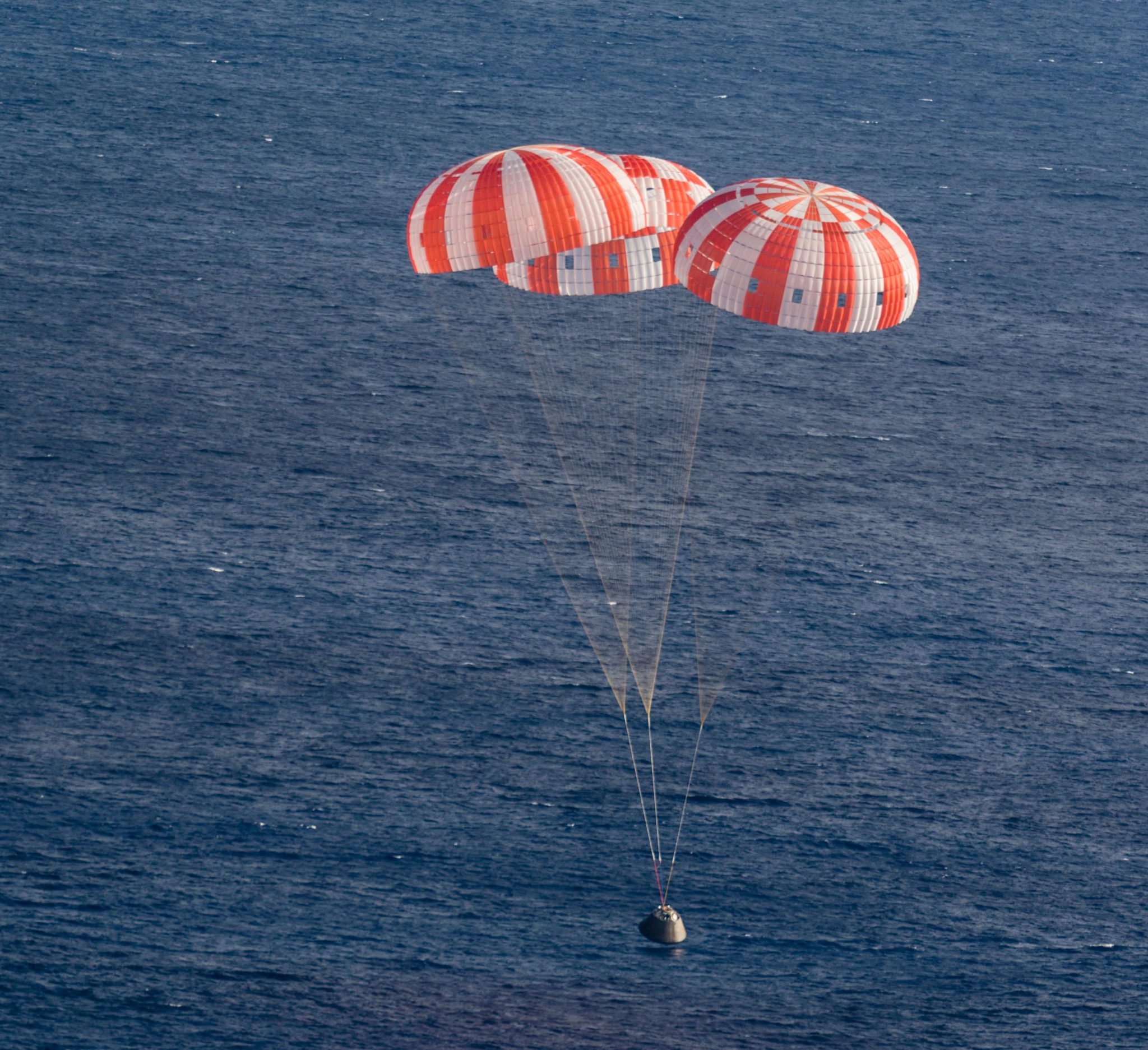 A white spacecraft in the shape of a capsule is hanging from two large parachutes that are red and white in color. The spacecraft is about to splash into the blue ocean.