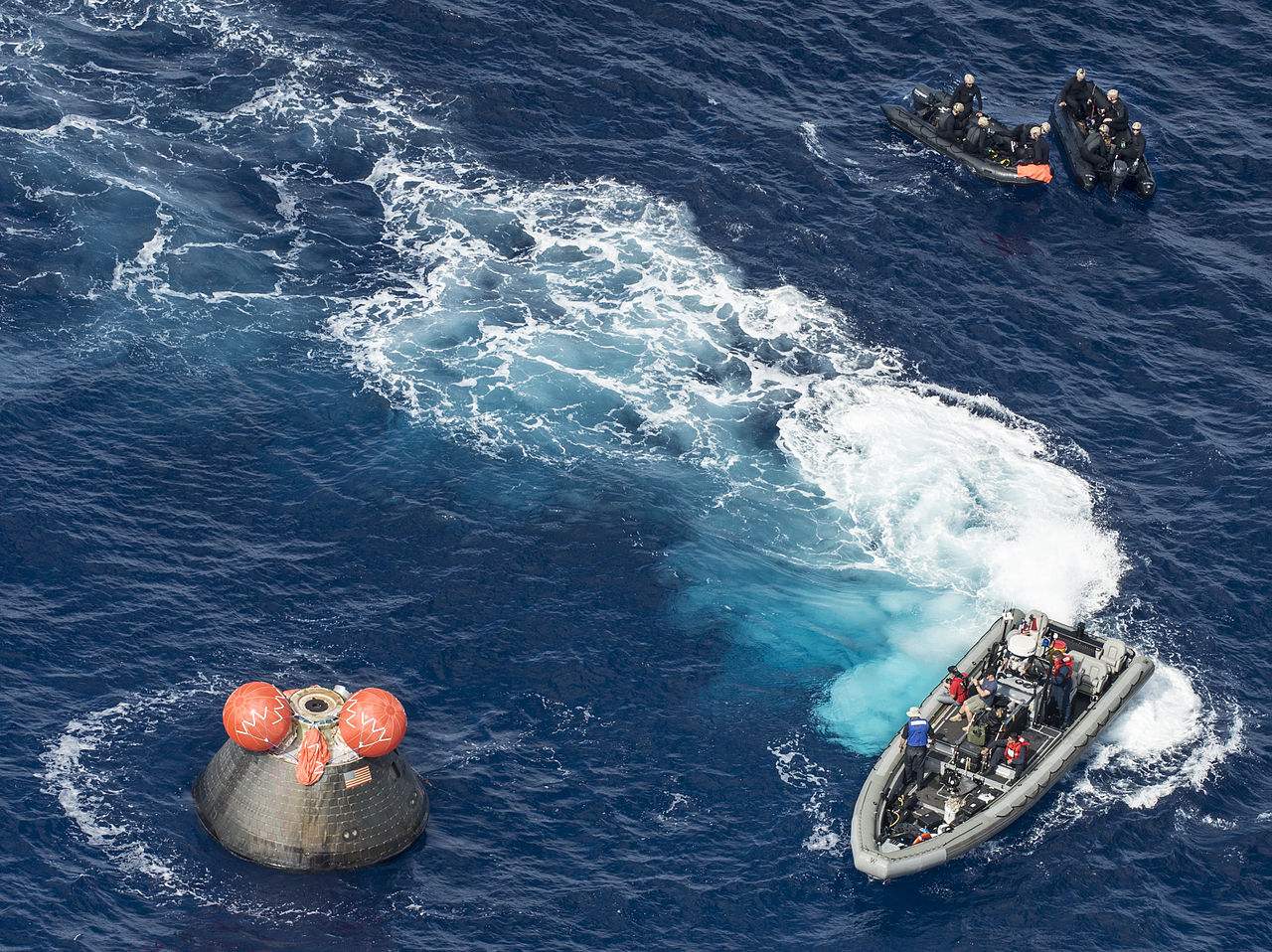 People in a high-speed boat are approaching a grey spacecraft shaped like a capsule that is on the water. The capsule has an American flag on it and inflated orange balloons on the top. Two other smaller boats are behind the first boat on standby. The boats are there to help the crew in the grey capsule exit the spacecraft.