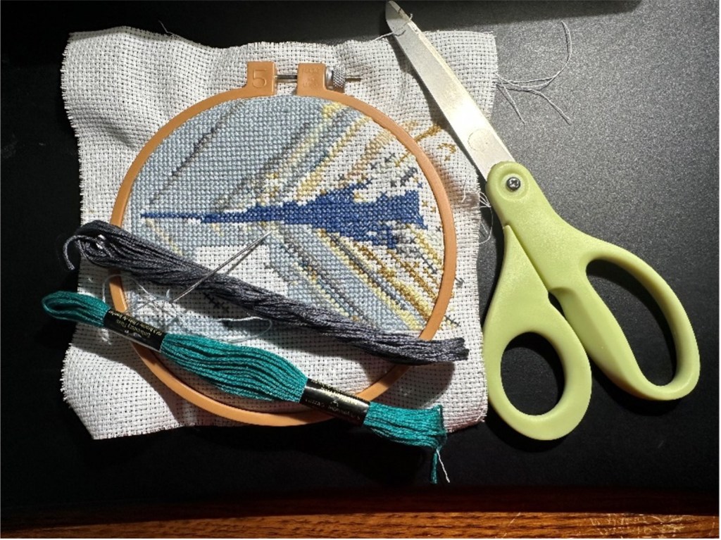 Cross-stitch of the Schlieren image of the X-59 in a hoop with scissors and two floss threads, in teal and gray.