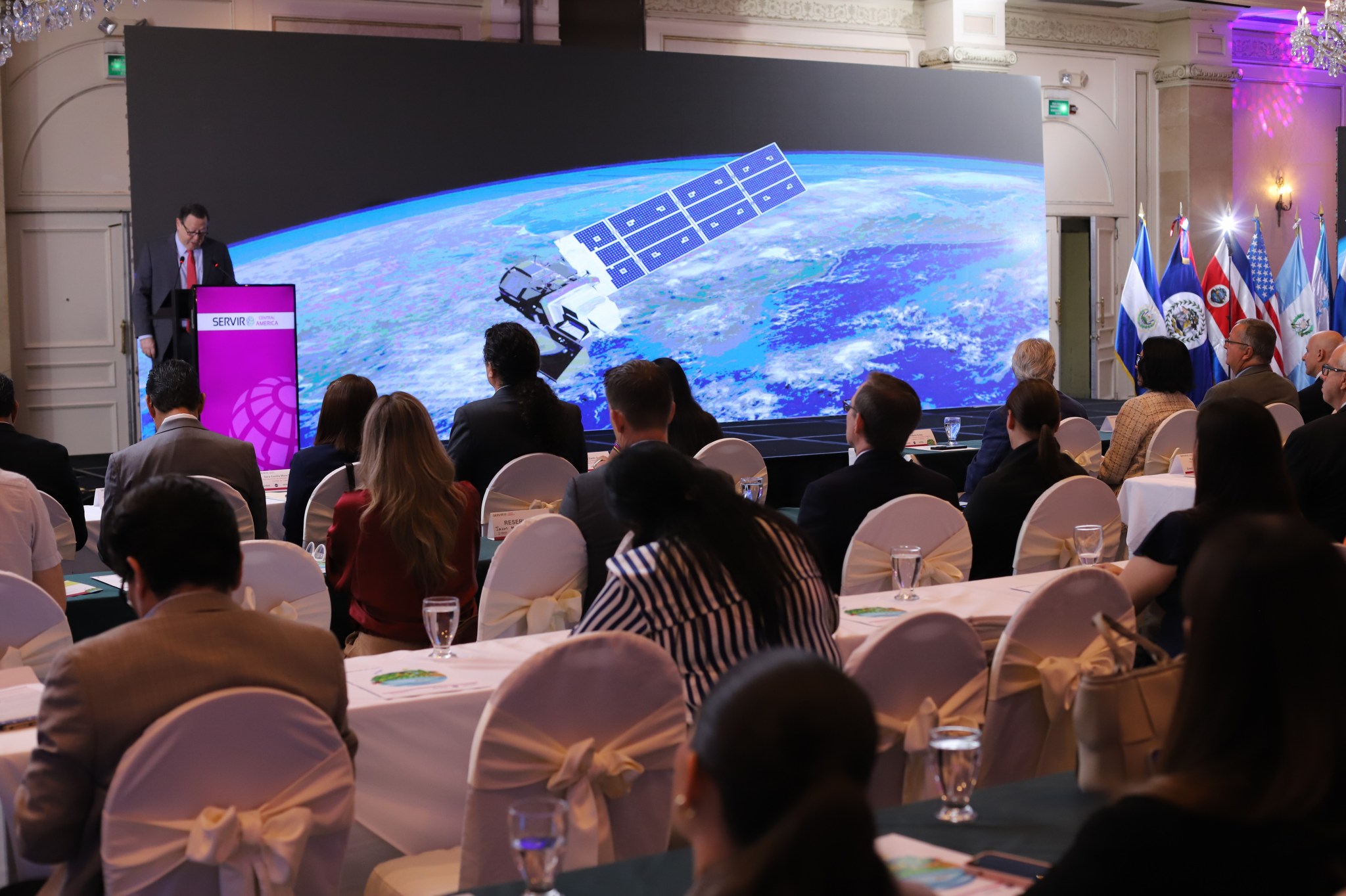 A group of people in a conference room sit in chairs looking at the speaker with the image of the Earth from space behind them.