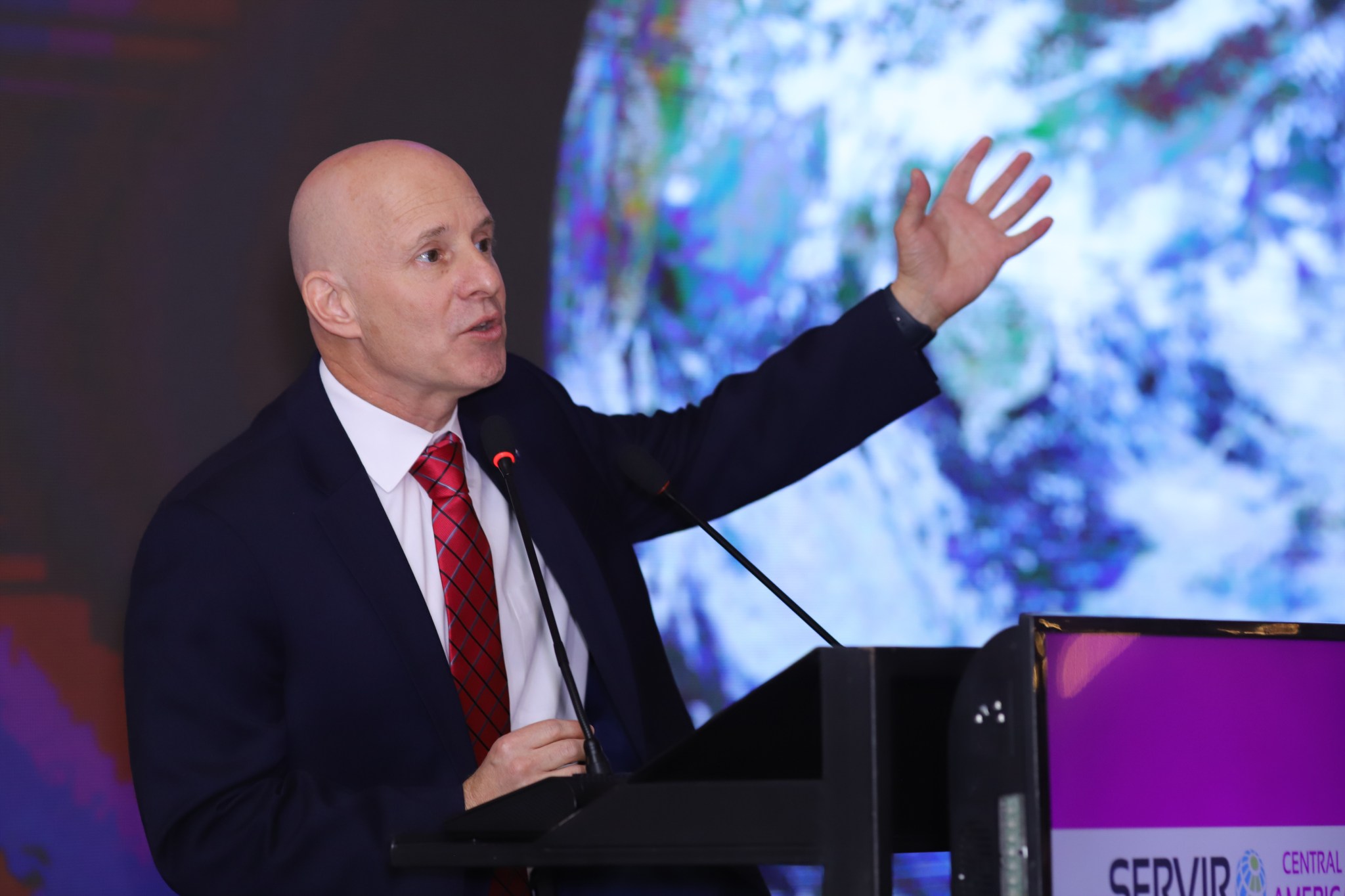 An older bald man with a black suit and red tie gestures at the photo of the Earth behind him.