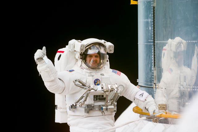 An astronauts wearing a spacesuit during a spacewalk gives a thumbs up, floating next to the Hubble Space Telescope