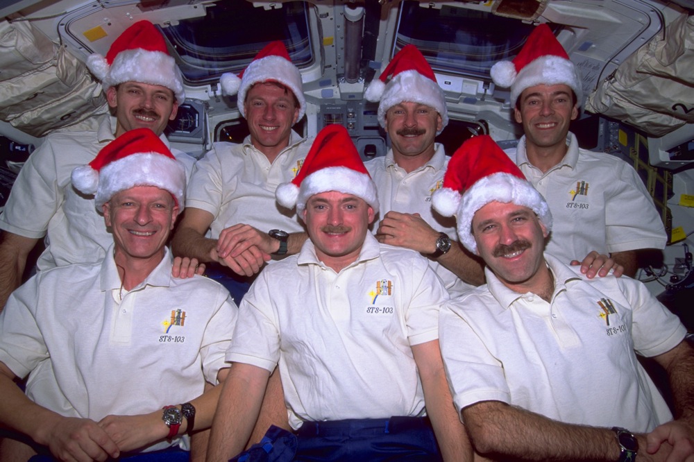 A group of seven astronauts wearing white shirts and Santa hats pose for a group photo