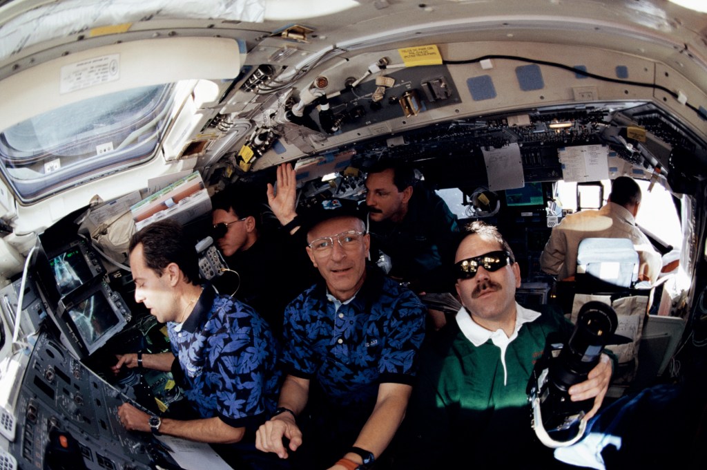 A fisheye lens view of a group of men crowded around the shuttle middeck, looking out the windows