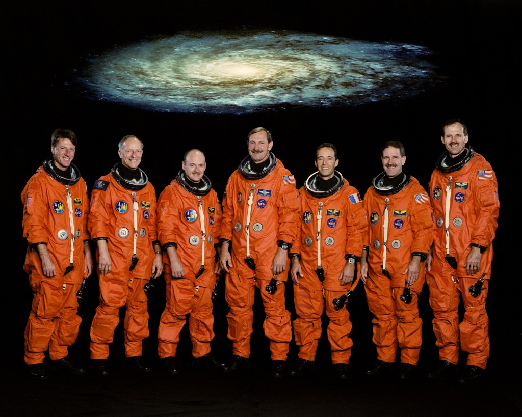 A group of 7 astronauts dressed in orange launch and entry suits with an image of a spiral galaxy above them