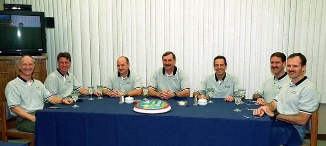 A group of 7 men seated at a long table, around a cake