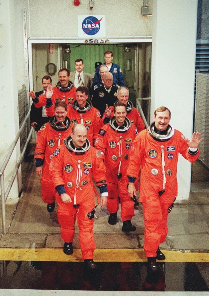 A group of 7 men all dressed in orange flight suits walk out of a building, waving